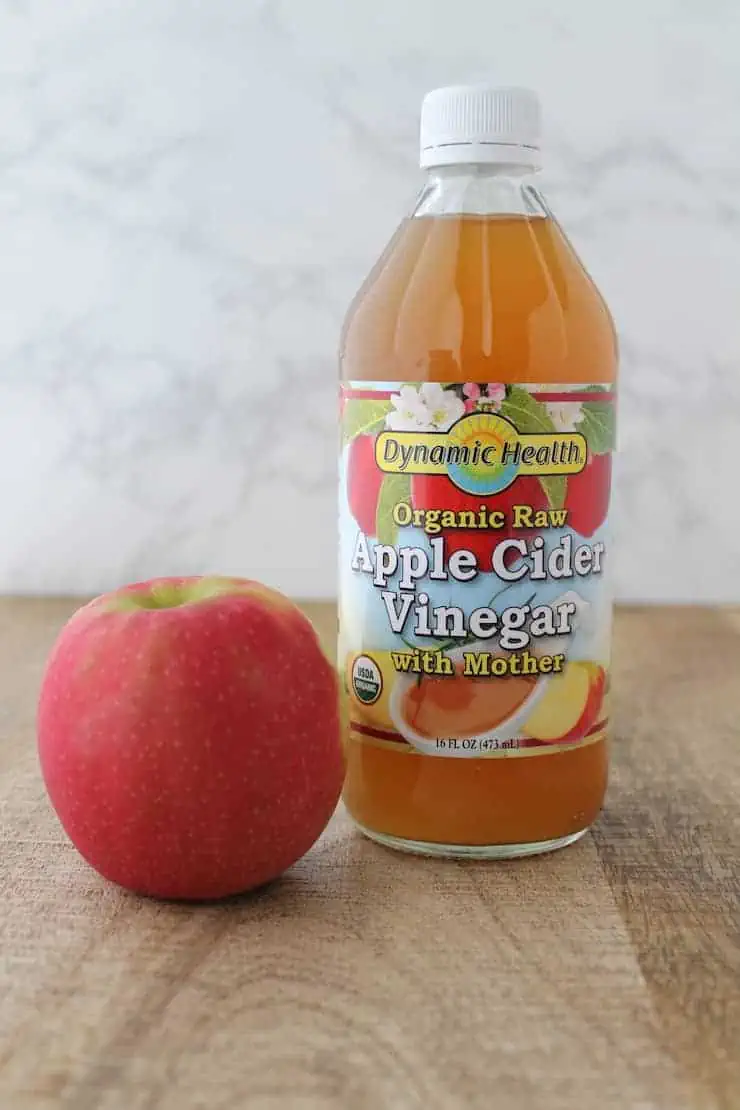 Glass bottle of apple cider vinegar on a wooden table next to a pink apple