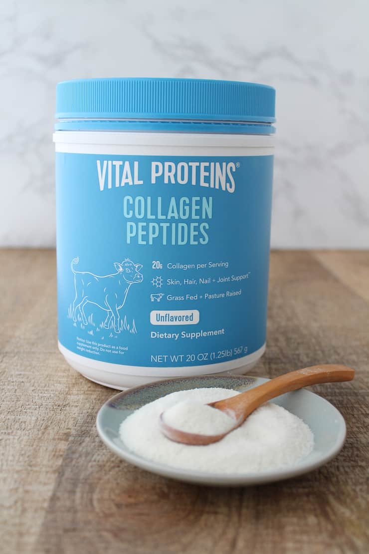 Blue and white tub of vital proteins collagen peptides on a wooden table next to a plate of collagen powder with a wooden spoon in it