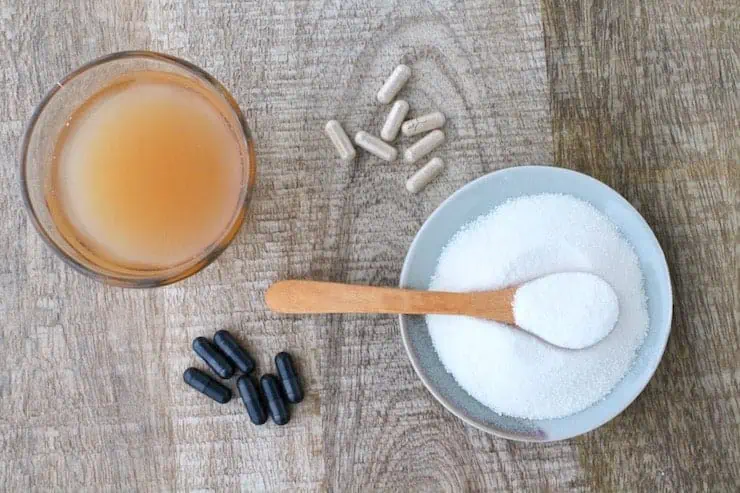 Plate of white powder with a wooden spoon in it next to black and brown supplement capsules and a glass of light brown liquid on a wooden table