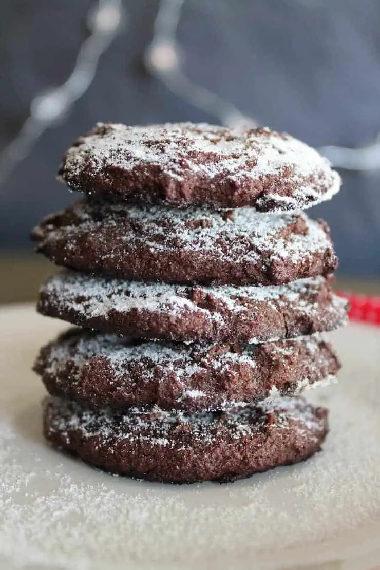 Stack of 5 peppermint keto chocolate cookies dusted with powdered sugar on white plate with a blue grey background and a red dish towel behind it
