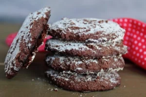 A stack of 4 peppermint keto chocolate cookies dusted with powdered sugar with 1 cookie leaning against the stack on a dark wooden table with a red dish towel with white polka dots behind it