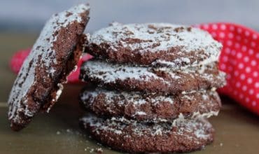 A stack of 4 peppermint keto chocolate cookies dusted with powdered sugar with 1 cookie leaning against the stack on a dark wooden table with a red dish towel with white polka dots behind it