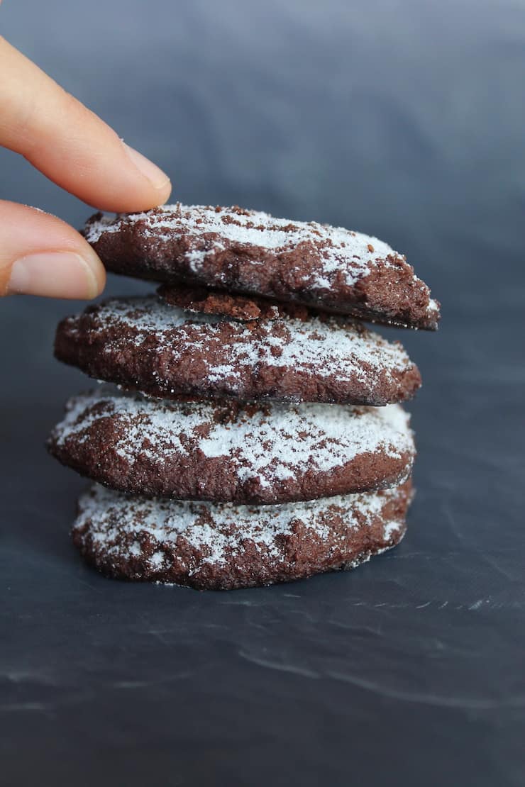 Stack of 4 peppermint keto chocolate cookies dusted with powdered sugar on a slate table with a hand reaching for a cookie