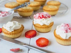 Yellow cupcake with white frosting on top with strawberry puree on top on white surface with cupcakes in the background and strawberry slices next to it