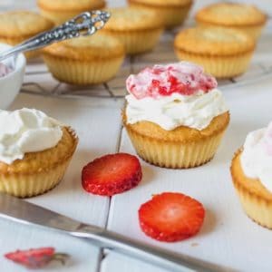 Yellow cupcake with white frosting on top with strawberry puree on top on white surface with cupcakes in the background and strawberry slices next to it