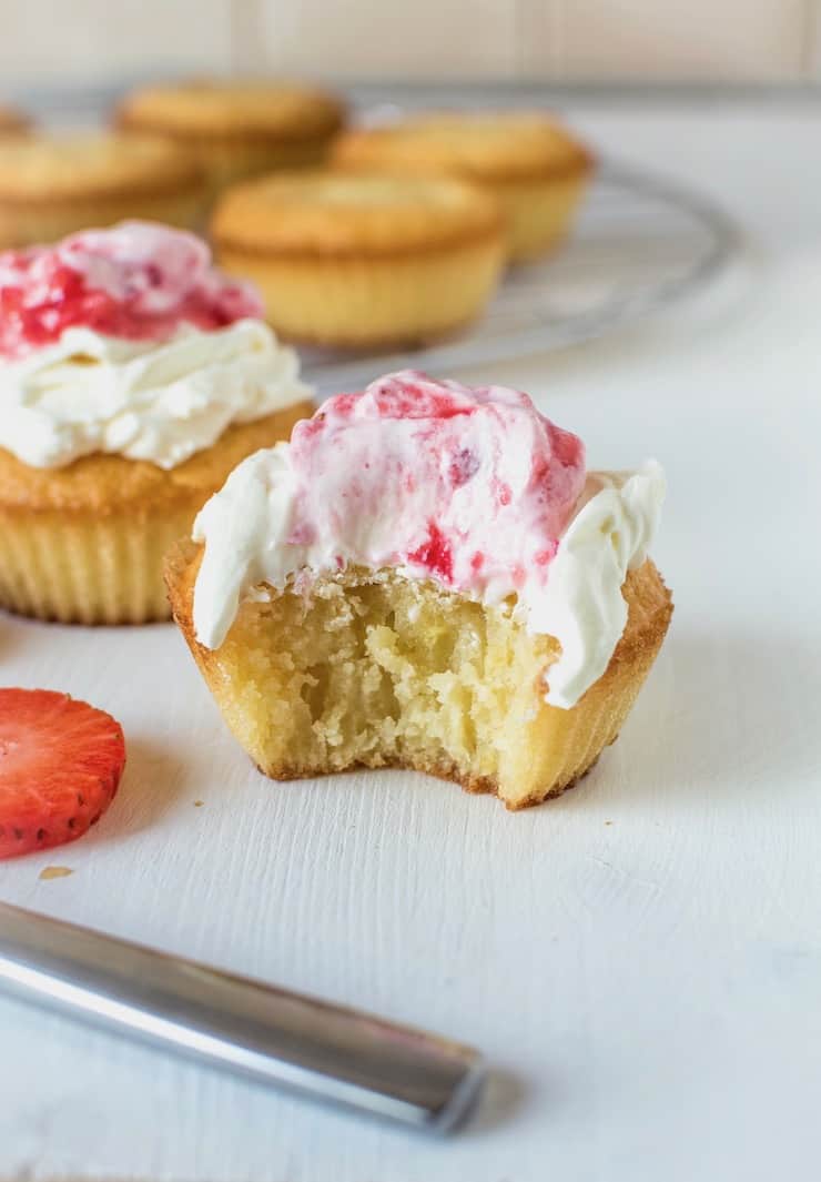 Close up of yellow cupcake with a bite taken out of it with white frosting on top with strawberry puree on top on white surface with cupcakes in the background and strawberry slices next to it