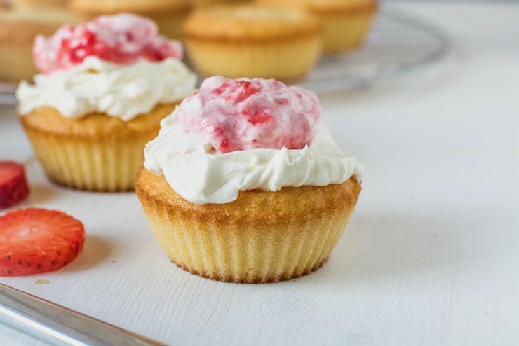 Yellow cupcake with white frosting on top with strawberry puree on top on white surface with cupcakes in the background and strawberry slices next to it