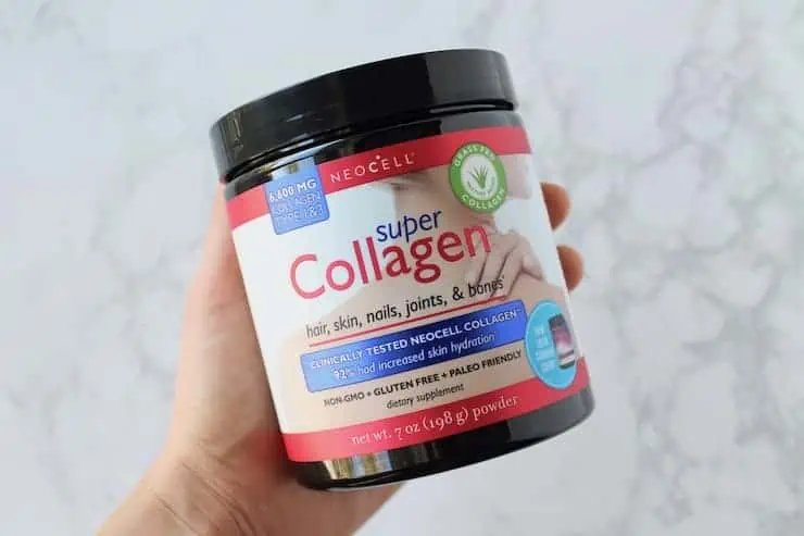 Hand holding a tub of collagen powder over a white marble surface