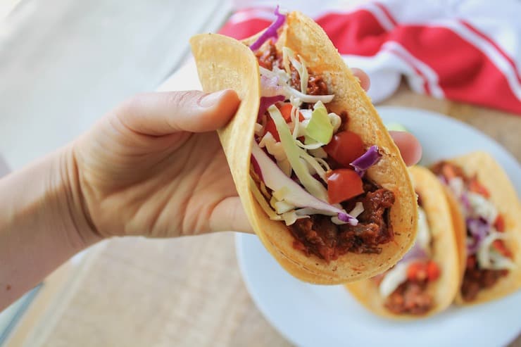 A hand holding one shredded beef taco with a plate of two tacos and a red an white dish towel on a wooden table in the background