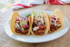 three shredded beef tacos on a white plate on a wooden table with a red and white dish towel in the background