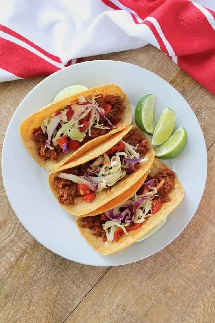 three shredded beef tacos on a white plate with lime slices on the plate on a wooden table with a red and white dish towel next to it