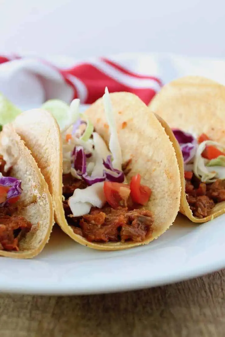 three shredded beef tacos on a white plate on a wooden table with a red and white dish towel in the background