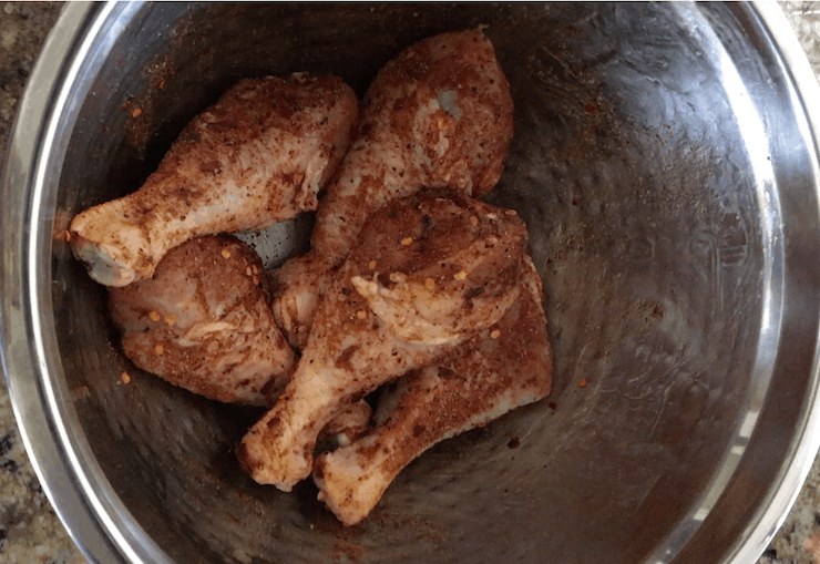 Raw chicken drumsticks in metal bowl covered in oil and spices