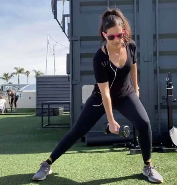 Woman in black workout clothes doing a side lunge with a weight in one hand on green grass