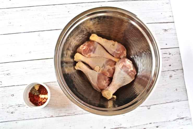 Metal bowl with 5 raw chicken drumsticks on a white wooden table with a small ramekin of spices next to it