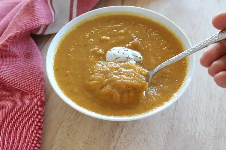 White bowl filled with orange pureed butternut squash soup with a spoon inside it and a dollop of yogurt in the middle on a wooden table with a red dish towel next to it