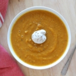 Overhead shot of white bowl filled with orange pureed butternut squash soup with dollop of yogurt in the middle on a wooden table with a red dish towel and spoon on either side