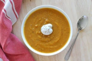 Overhead shot of white bowl filled with orange pureed butternut squash soup with dollop of yogurt in the middle on a wooden table with a red dish towel and spoon on either side