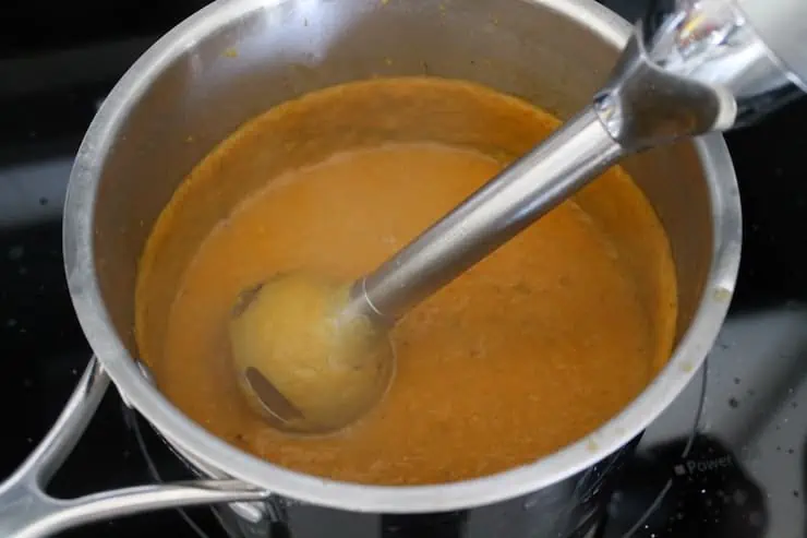 Pot on the stove filled with pureed butternut squash soup with immersion blender in it