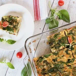 Square slice of turkey veggie baked frittata on a white plate on a white wooden surface with cherry tomatoes and basil next to it with the full frittata dish next to it