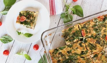 Square slice of turkey veggie baked frittata on a white plate on a white wooden surface with cherry tomatoes and basil next to it with the full frittata dish next to it