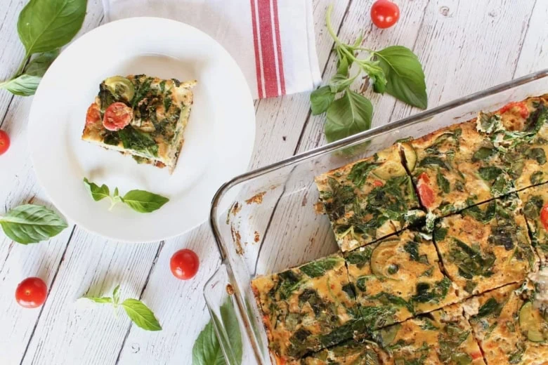 Square slice of turkey veggie baked frittata on a white plate on a white wooden surface with cherry tomatoes and basil next to it with the full frittata dish next to it