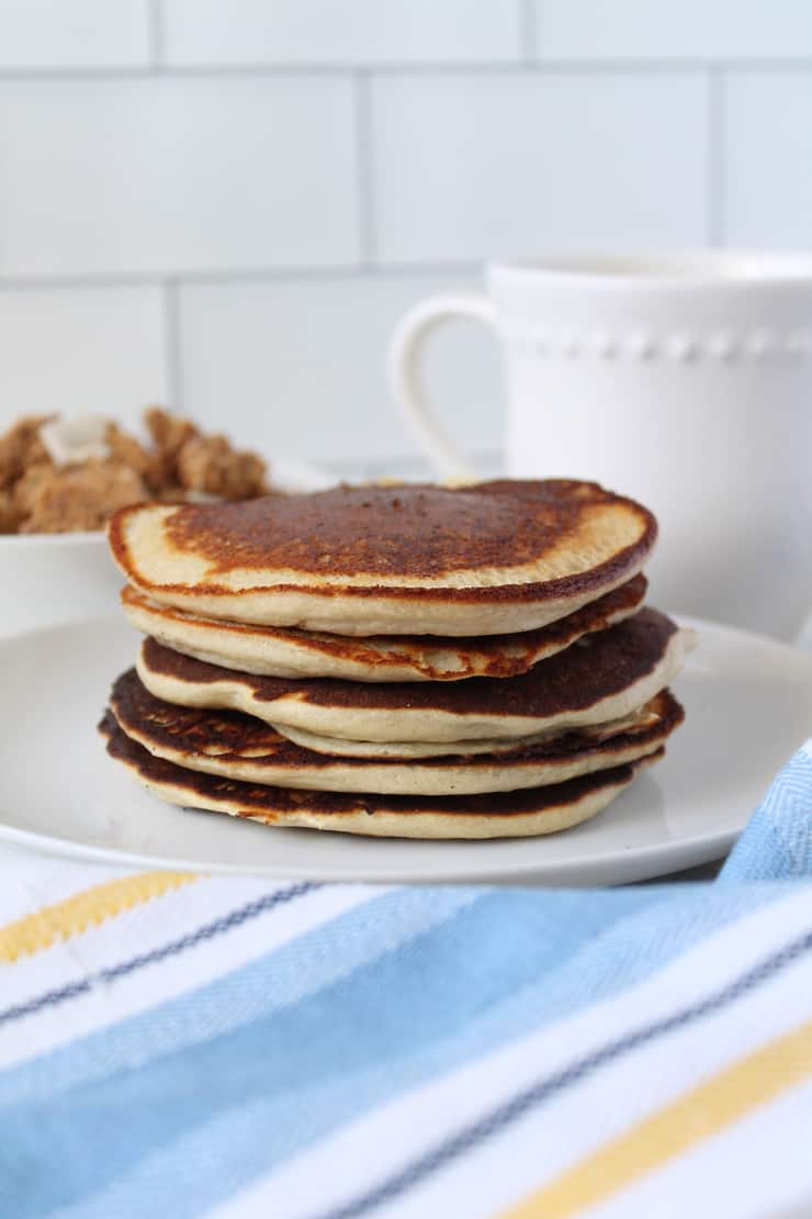 Close up of stacked pancakes on a white plate with a blue, white and yellow striped kitchen towel next to it with a white bowl filled with granola and a white mug in the background on a white marble surface