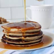 Close up of stacked pancakes with maple syrup being poured over them on a white plate with a blue, white and yellow striped kitchen towel next to it with a white bowl filled with granola and a white mug in the background on a white marble surface