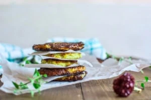 gluten free zucchini fritters stacked on top of each other with parchment paper in between on a brown wooden table with green herbs next to it