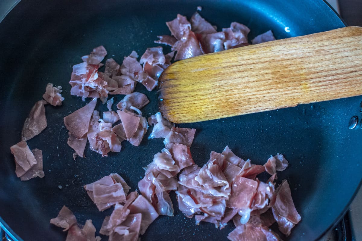 prosciutto frying in a black cast iron skillet with a wooden spatula mixing it