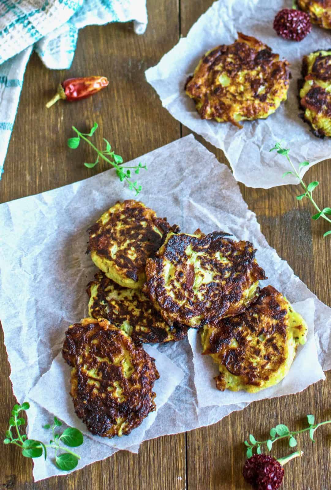 several zucchini fritters laid out on white parchment paper on a brown wooden table with green herbs next to it