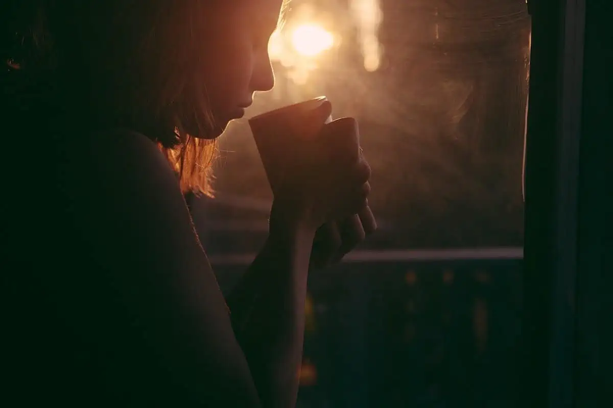 silhouette of a woman drinking a mug of tea with sun at dusk shining through a window