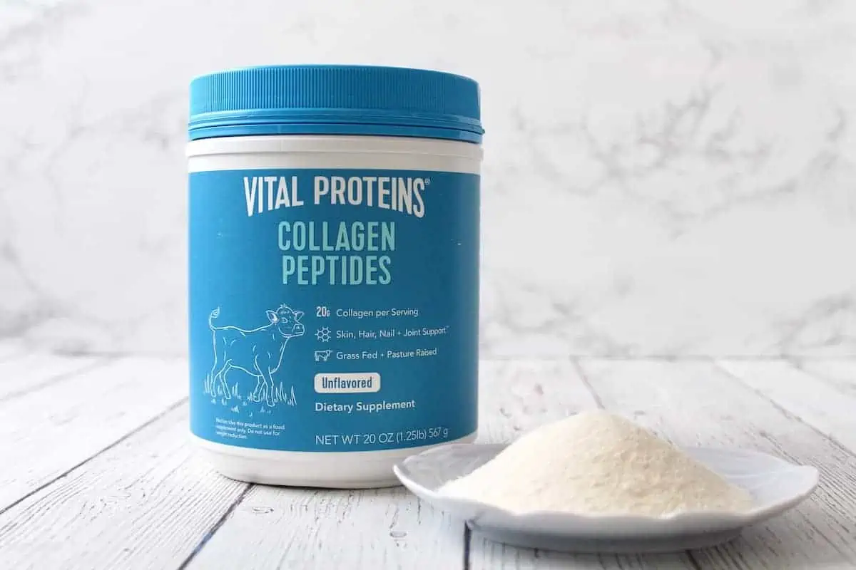 Blue tub of vital proteins collagen peptides next to dish with white powder in it on a white wooden surface with marble background