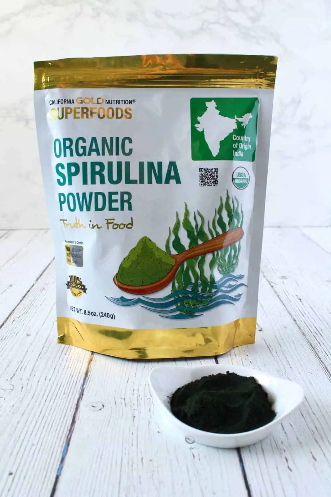 White and gold bag of organic spirulina powder next to a small white dish with dark green powder in it on a white wooden surface with a white marble background