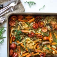 Sheet pan filled with herb roasted chicken and vegetables on a white wooden table next to a napkin with knives and forks on it next to herb sprigs