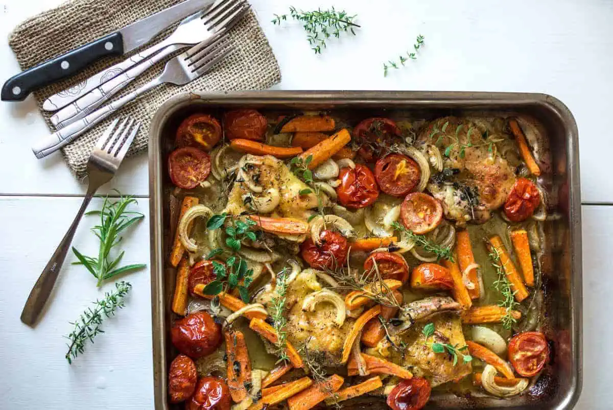 Sheet pan filled with herb roasted chicken and vegetables on a white wooden table next to a napkin with knives and forks on it next to herb sprigs