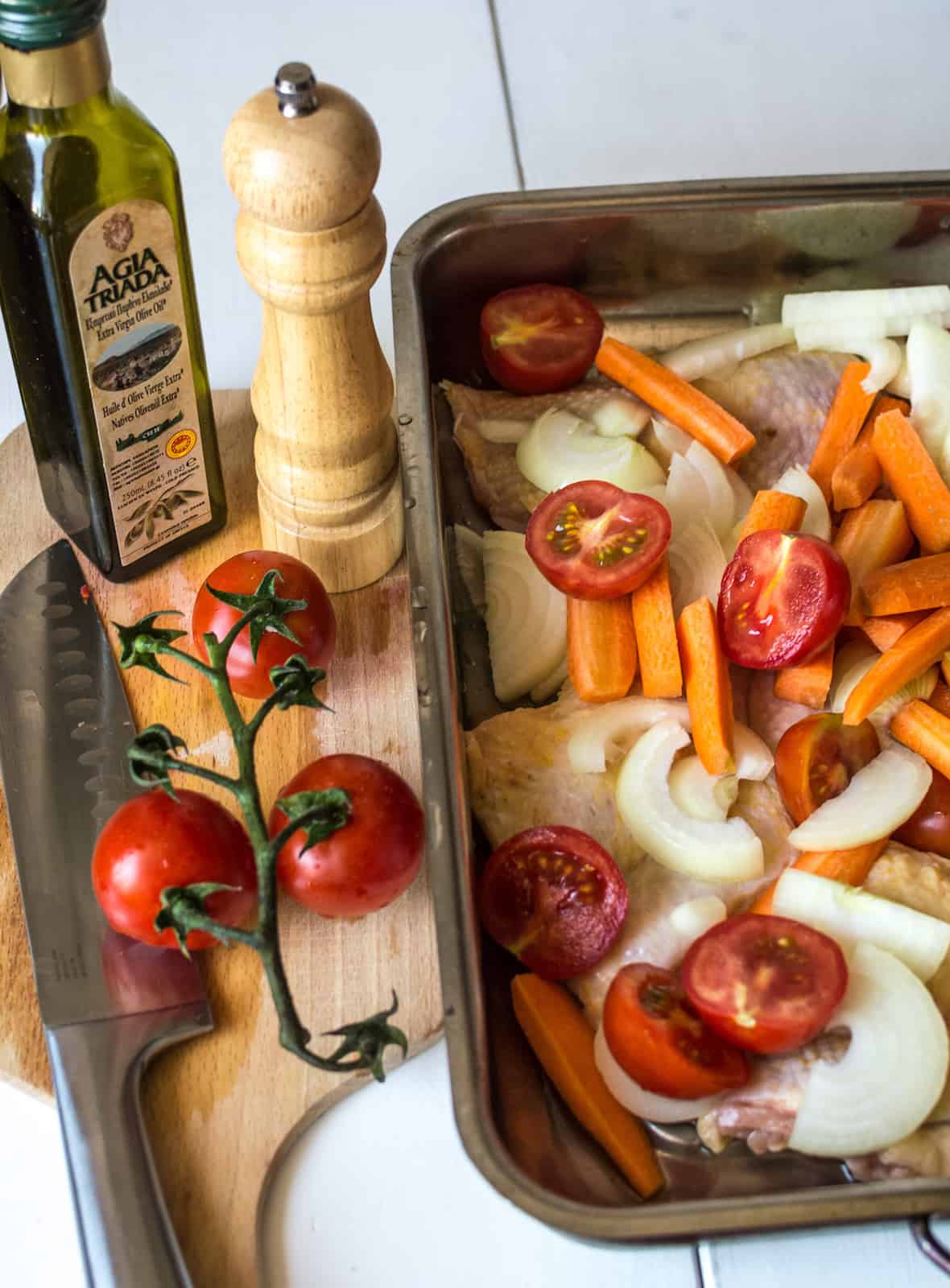 Uncooked chicken and vegetables in a sheet pan next to a cutting board with a chefs knife, tomatoes, olive oil and a pepper grinder on it
