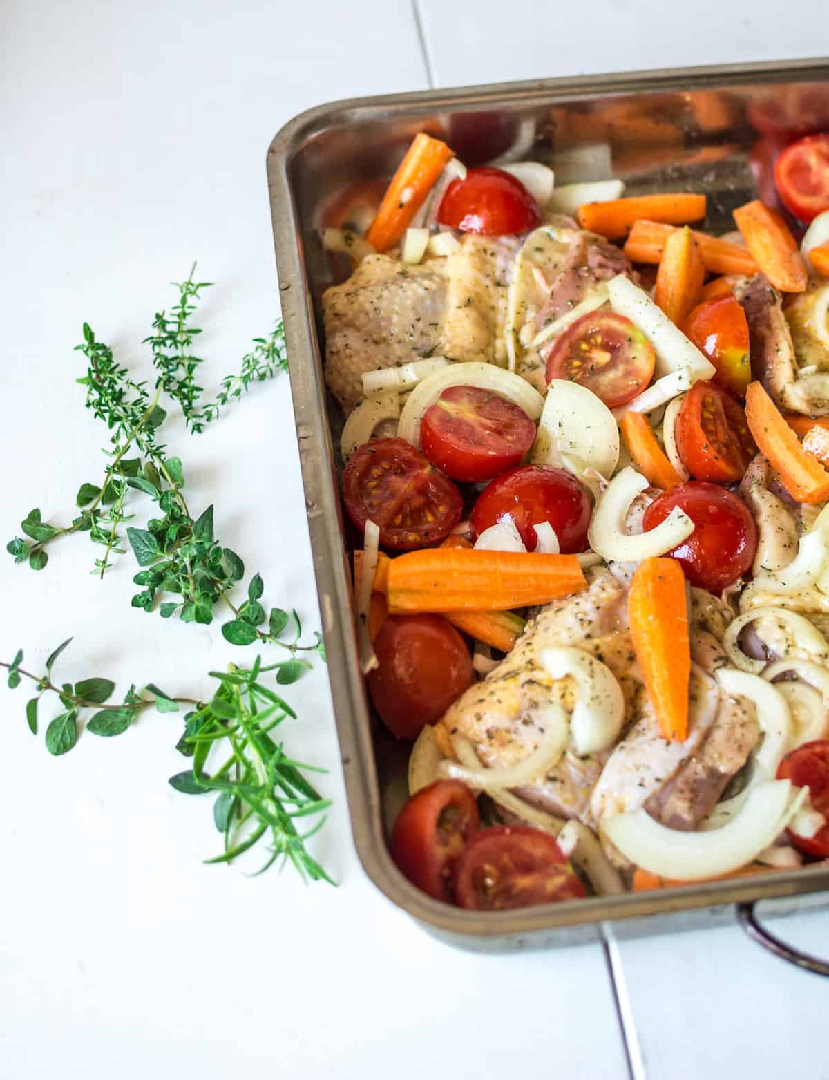 Sheet pan with uncooked chicken and vegetables in it sprinkled with spices and herbs on a white wooden table with herb sprigs next to it