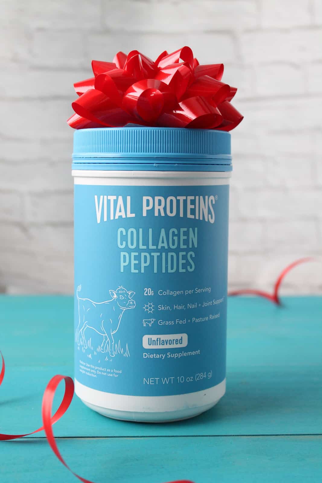 blue tub of vital proteins collagen with a red bow on top next to a red ribbon on a teal wooden table with a white brick background