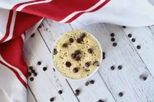 Finished chocolate chip mug cake in a small ramekin on a white wooden surface with chocolate chips surrounding it and a red and white striped dish cloth next to it