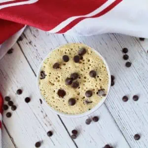 Finished chocolate chip mug cake in a small ramekin on a white wooden surface with chocolate chips surrounding it and a red and white striped dish cloth next to it