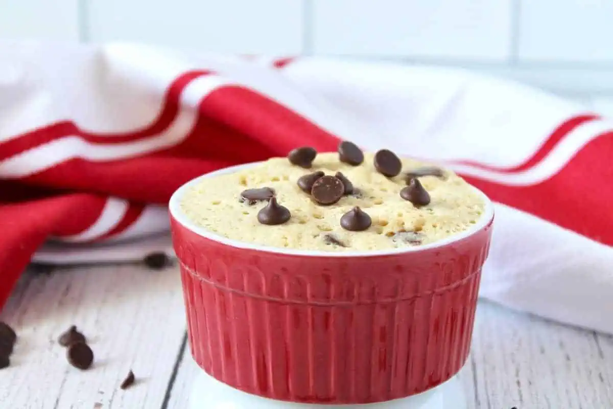 Finished chocolate chip mug cake in a red ramekin on a white wooden surface with a red and white striped dish cloth in the background
