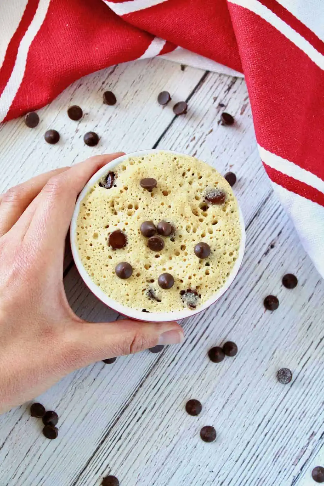 Hand holding a finished chocolate chip mug cake in a small ramekin on a white wooden surface with chocolate chips surrounding it and a red and white striped dish cloth next to it