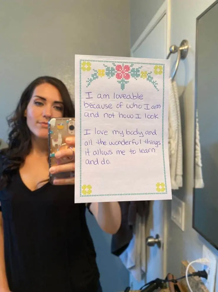 Paper stuck to bathroom mirror with affirmations written on it with woman in the background of the mirror taking picture with phone