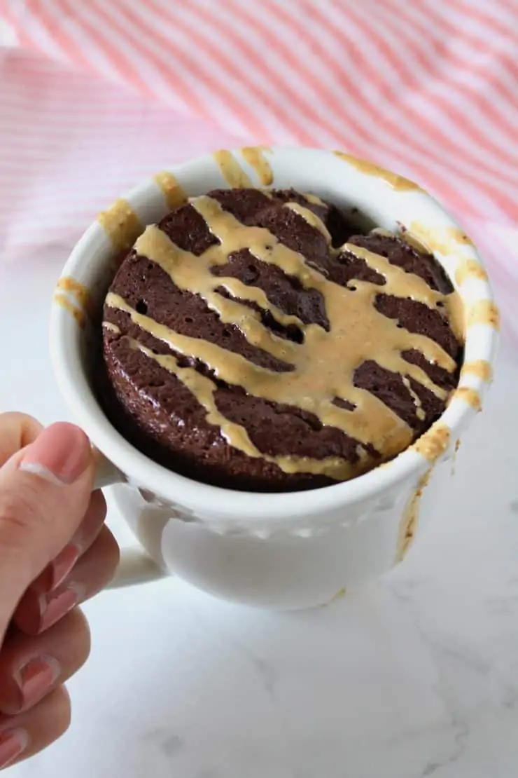 Hand holding a white mug filled with chocolate cake drizzled with peanut butter over a white marble surface next to a pink and white striped dish towel