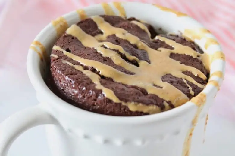 Close up shot of a white mug filled with chocolate cake drizzled with peanut butter on a white marble surface next to a pink and white striped dish towel