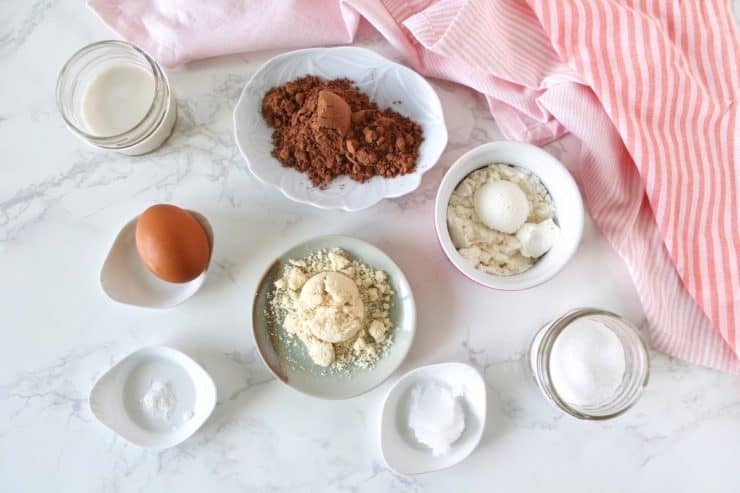 Overhead shot of protein mug cake ingredients in various dishes on a white marble surface next to a pink and white striped dish towel