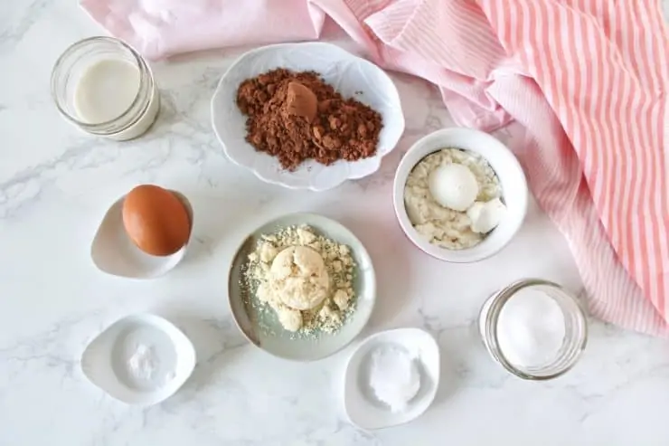 Overhead shot of protein mug cake ingredients in various dishes on a white marble surface next to a pink and white striped dish towel