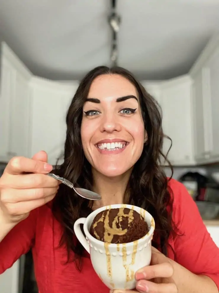 Smiling dark haired woman with red shirt on holding mug cake with spoon in other hand