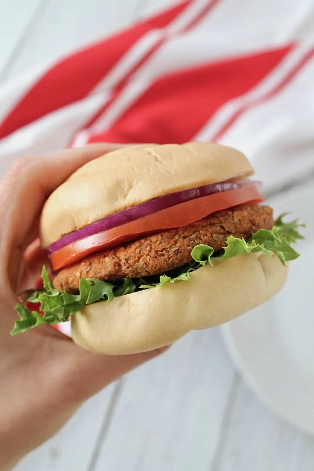 Close up of hand holding a lentil burger with lettuce, tomato, onion and a bun with a red and white striped dish towel in the background